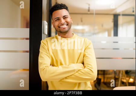 Smiling indian businessman standing in modern open space office and looking at the camera. Young positive male student, proud and successful multiracial small business owner Stock Photo