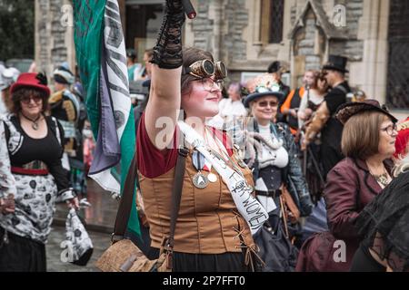 A group of steampunks walking in procession along a street. A young woman wearing a sash that says Votes for Women waving a flag. Stock Photo