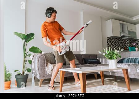 Caucasian man wearing wireless headphones plays rocks air guitar on vacuum cleaner. Male doing home chores with fun Stock Photo