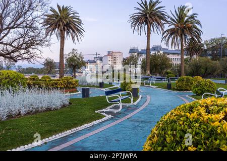 Palm trees, bushes, paths and benches in a public park Jardines de Piquio at sunset, with the city view in the background. Santander, Cantabria, Spain Stock Photo