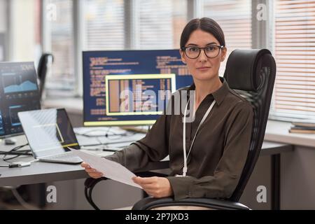 Portrait of woman wearing glasses looking at camera while sitting at desk in IT development office with computers in background Stock Photo