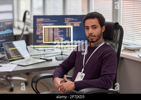 Portrait of male data scientist looking at camera while sitting at desk in IT development office with computers in background Stock Photo