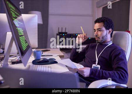 Side view portrait of IT developer working at night writing code and holding documents at desk in office Stock Photo