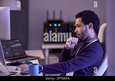 Side view portrait of software developer working at night writing code at desk in office lit by neon lights Stock Photo