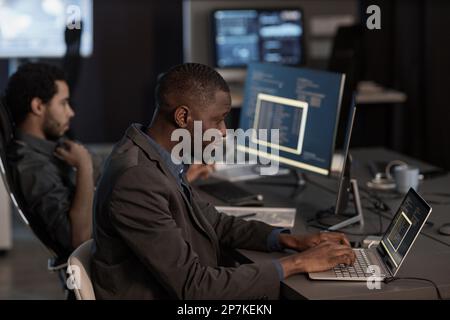Side view portrait of black man writing code and typing at laptop keyboard while working in data security office Stock Photo