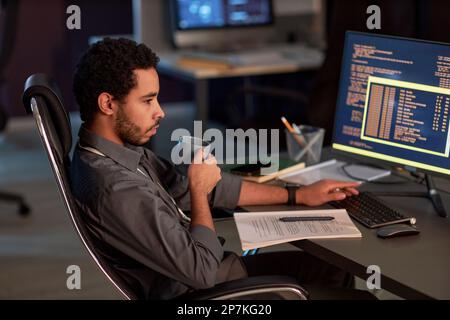 Side view portrait of male software developer writing code and drinking coffee while working at night in data security office Stock Photo