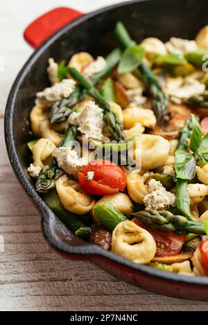 Cast iron pan with vegetable tortellini with asparagus, tomatoes, mushrooms and home made vegan feta cheese. Stock Photo