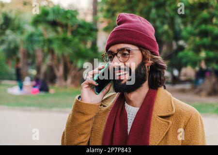 One bearded happy male with eye glasses and beanie hat having a cell phone conversation standing at park outdoors. Close up portrait of a guy smiling and talking by smartphone. High quality photo Stock Photo