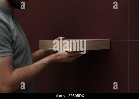 Man using cross line laser level for hanging shelf on brown wall, closeup Stock Photo