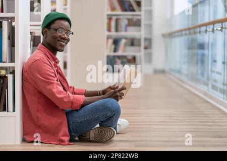 Smiling Black student man reading book in university library , looking at camera. Reading hobby.  Stock Photo