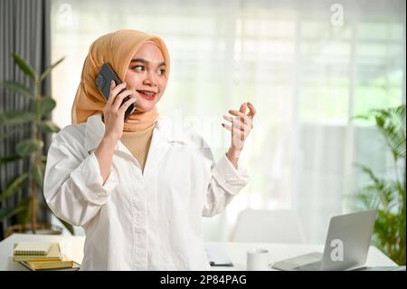 Confident and professional millennial Asian Muslim businesswoman or female marketing manager talking on the phone with her business client in her offi Stock Photo