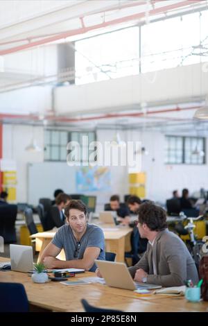 Exchanging ideas in the office. two colleagues talking together while working on laptops at a desk in a large office. Stock Photo