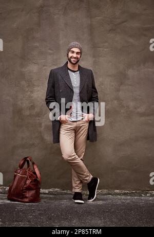 Ive got all the time in the world. a handsome young man leaning against a wall outside in the city with his bag waiting for someone. Stock Photo