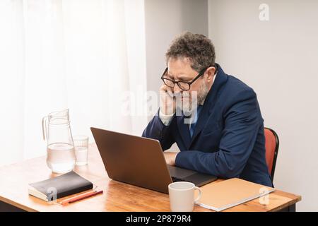 Experienced businessman in his 50s, wearing a suit and tie, intently reviewing an important document on his computer at his desk Stock Photo