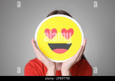 Woman covering face with heart eyes emoji on grey background Stock Photo