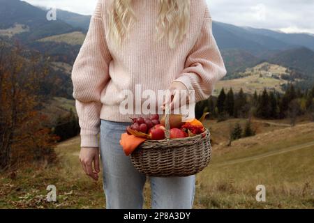 Woman holding wicker picnic basket with fruits and autumn leaves in mountains, closeup Stock Photo