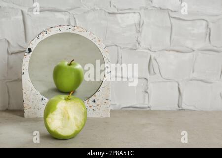 Bitten green apple near mirror with reflection of whole fruit on table, space for text Stock Photo