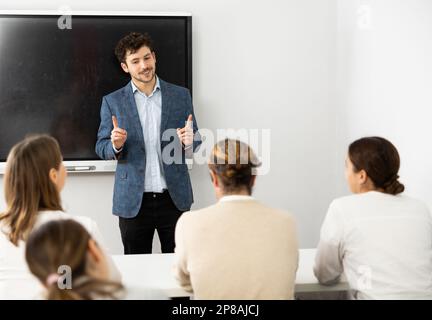 Male business coach standing near interactive board explaining topic while standing against group of people sitting at desks in front of him Stock Photo