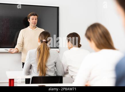 Male business coach explaining topic while standing against group of people sitting at desks in front of him Stock Photo