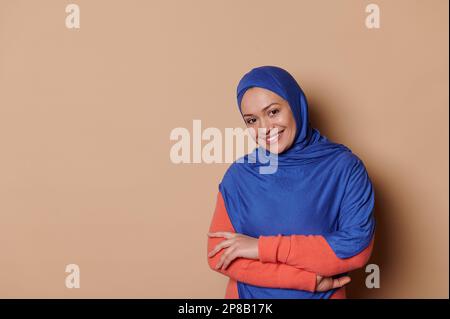 Arab Muslim woman in blue hijab, smiling looking at camera, beige background. Strict formal outfit. Elegant appearance. Stock Photo