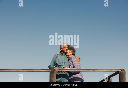 Romantic elderly couple smiling and embracing each other while standing on a wooden foot bridge. Happy senior couple enjoying a relaxing seaside holid Stock Photo