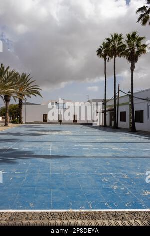 Blue playground of a school basketball stadium, lined by high palm trees. Surrounded by white buildings. Cloudy rainy sky in the winter. Casillas del Stock Photo