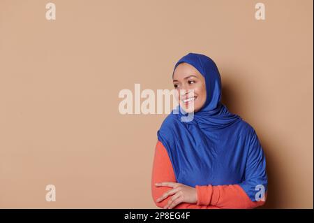 Arab Muslim woman in blue hijab, smiling looking aside at ad space on beige background. Islamic female fashion. Elegance Stock Photo