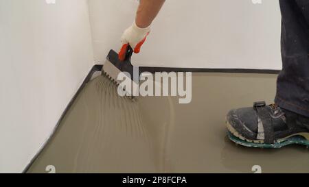 Repairman in protective gloves and uniform using a metal spatula to level a liquid self-levelling floor screed. A worker pours liquid cement mortar at Stock Photo