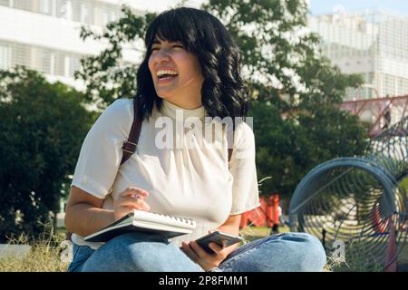 young venezuelan woman sitting in the park at the university, she is excited, laughing out loud, she is distracted by jokes she saw on social networks Stock Photo