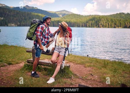 young caucasian adult couple taking a break in nature, looking each other, smiling, leaned on a wooden bench. active, healthy lifestyle concept Stock Photo