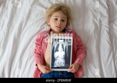 Toddler child, blond boy, holding x-ray picture on tablet of child body with swallowed magnet showing, child swallow dangerous object Stock Photo