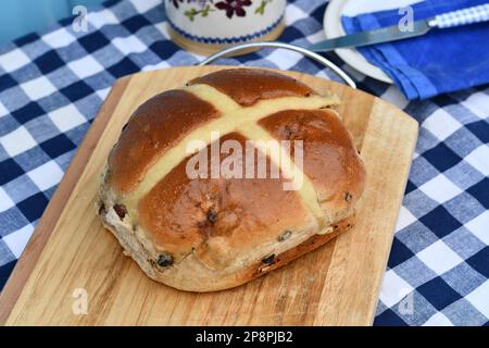 Giant hot cross bun on a chopping board. Traditional spiced, sticky glazed fruit buns with pastry crosses. Served as a classic Easter treat. Stock Photo