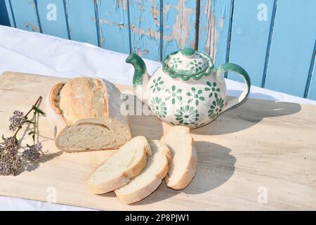 Sliced loaf of bread on a cutting board with a blue wooden background with a green and white coloured teapot Stock Photo