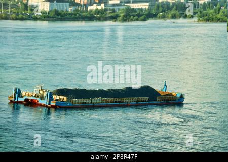 A river non-propelled barge barge (side stone dump vessel) with rubble is anchored in the middle of the river waiting for a tug Stock Photo