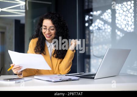 Successful woman financier accountant reading company financial report rejoices and celebrates victory and good achievement results, businesswoman inside office paper work using laptop at workplace. Stock Photo