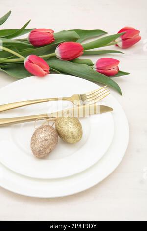 Easter table setting, Red tulips and Easter eggs decoration, golden cutlery on plates, white background Stock Photo