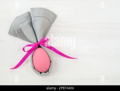 Easter table setting, Pink Easter egg in a gray napkin, rabbit ears shape, on white wooden background, top view Stock Photo