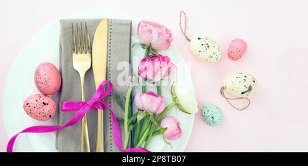 Easter table setting, Easter eggs and flowers decoration, napkin and golden cutlery on white plates, pastel pink background Stock Photo