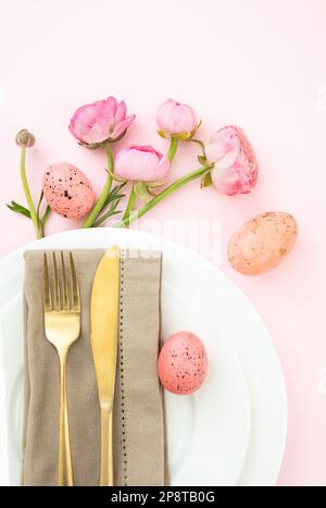 Easter table setting, Easter eggs and flowers decoration, napkin and golden cutlery on white plates, pastel pink background Stock Photo