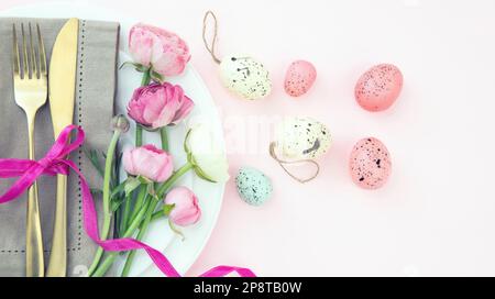 Easter table setting, Easter eggs and flowers decoration, napkin and golden cutlery on white plates, pastel pink background Stock Photo