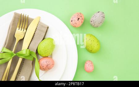 Easter table setting, Easter eggs decoration, napkin and golden cutlery on white plates, pastel green background Stock Photo