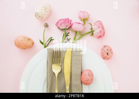 Easter table setting, Easter eggs and flowers decoration, napkin and golden cutlery on white plates, pastel pink background Stock Photo