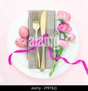 Easter table setting, Easter eggs and flowers decoration, napkin and golden cutlery on white plates, pastel pink background Stock Photo