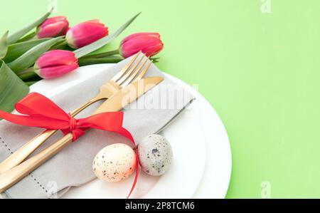 Easter table setting, Spring flowers and eggs decoration, napkin and golden cutlery on white plates, pastel green background Stock Photo