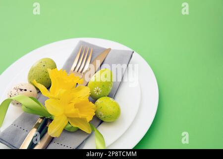 Easter table setting, Spring flowers and eggs decoration, napkin and golden cutlery on white plates, pastel green background Stock Photo