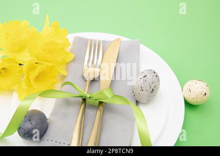 Easter table setting, Spring flowers and eggs decoration, napkin and golden cutlery on white plates, pastel green background Stock Photo