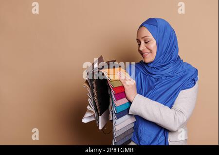 Smiling Muslim woman in blue hijab, putting her hand on the upholstery textile color swatches, isolated beige background Stock Photo