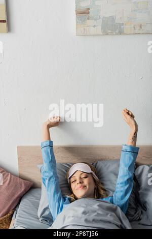 tattooed woman in sleeping mask and nightwear stretching while lying in bed,stock image Stock Photo