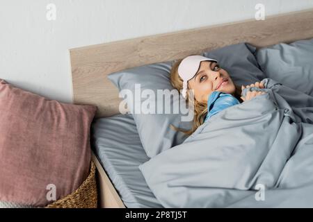 cheerful woman in sleeping mask lying under duvet in comfortable bed,stock image Stock Photo