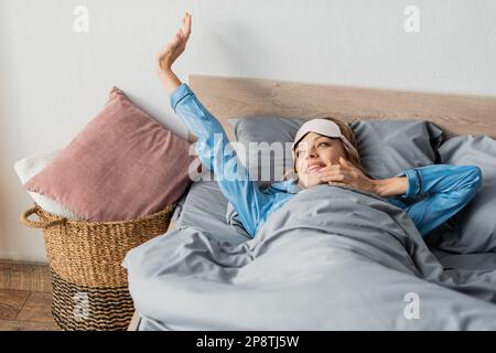 cheerful woman in sleeping mask and nightwear stretching while lying in bed,stock image Stock Photo